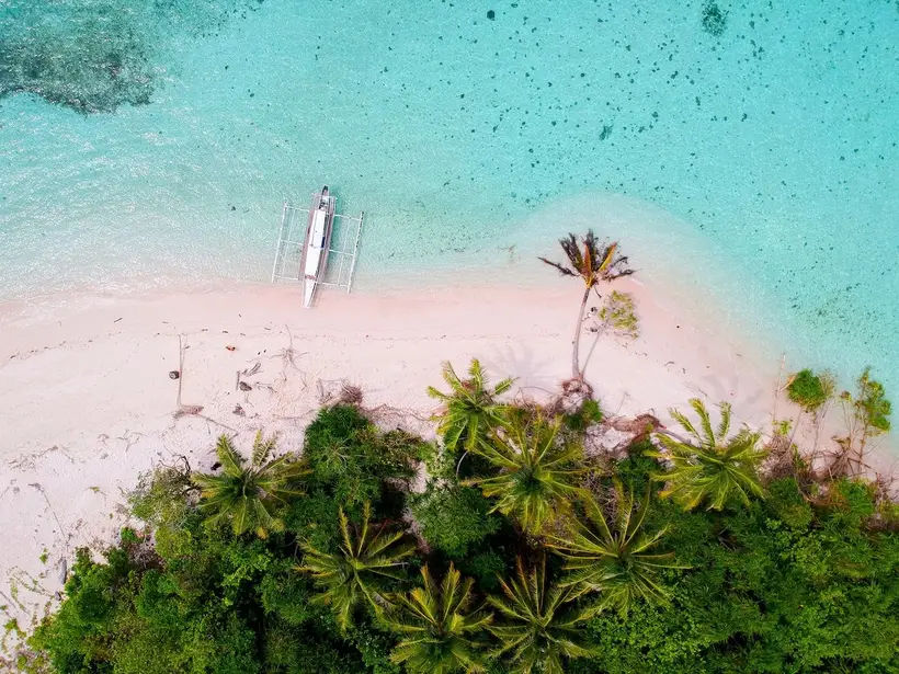 Aerial view of a pale pink sandbar, palms and a small outrigger boat in Balabac